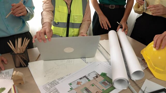 Architect and safety engineer discussion about building blueprint displaying on laptop on table with architectural document and wooden block scatter around at modern office. Closeup. Delineation.