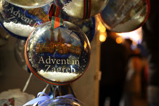 The image shows a close-up of a Christmas ornament with the text "Advent in Zagreb." The ornament features a depiction of a cathedral and is adorned with a red and green ribbon.