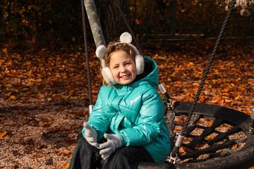 Smiling little girl on playground with headphones sitting on swing and laughing.