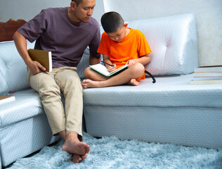 father and son are seated together on a comfortable sofa, collaborating on homework. The boy holds a book, while the father uses a textbook to complement their study session.education, family concept