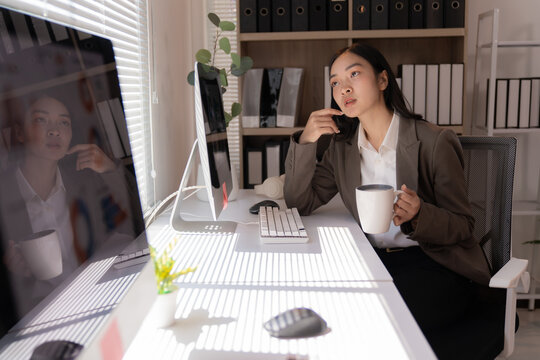 Young asian businesswoman drinking coffee and thinking about new business project while sitting at workplace in modern office, sunlight from window reflecting on desk