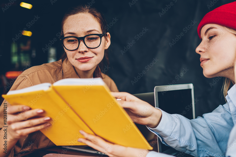 Poster hipster student in red hat showing textbook with useful information to colleague during collaboratio