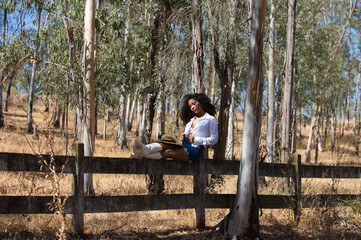 Fototapeta premium woman, young, beautiful, Latin, brunette with curly hair dressed in white shirt, denim skirt, white boots and hat, sitting with her legs stretched out on the forest fence. The woman rests.