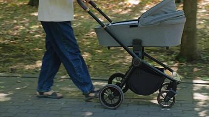 Parent pushing modern black stroller with bassinet along shaded brick pathway, wearing blue jeans with sandals, surrounded by trees and fallen leaves. A walk in the park with an infant in a pram