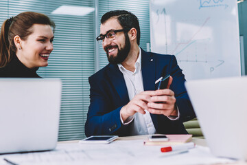 Young handsome smiling graphic designer with attractive female colleague watching funny video in...