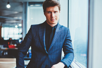 Fototapeta premium Portrait of confident businessman in elegant suit standing near office window and looking at camera, handsome caucasian male entrepreneur spending time at coworking space and waiting colleague