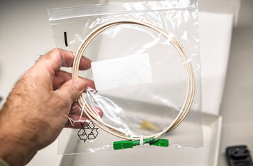 Hand holding a fiber optic cable sealed in a transparent plastic bag with green connectors,...