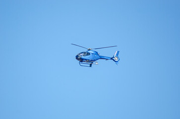 A small blue helicopter is flying through a clear sky. The helicopter has two long blades and appears to be either taking off or landing. It seems to be moving at a slow speed as it glides over a fiel