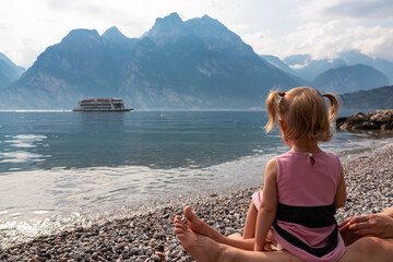 Toddler sits on legs of adult on pebble beach watching ferry boat passing in lakeside town...