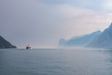 Ferry boat floating along coastline of misty Lake Garda covered with haze seen from Nago-Torbole,...