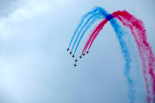 A stunning display of synchronized aerobatics featuring multiple airplanes leaving vibrant red and blue smoke trails in a dramatic V-shaped formation against a cloudy sky. Perfect for themes of teamwo