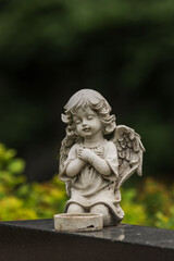 Fototapeta premium Small angel statue on a tombstone. Small-sized grave decoration angel at the cemetery in Finland. Lutheran cemetery in a beautiful park-like landscape. Empty space for text.