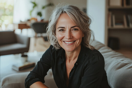 Mujer madura de 40 a&ntilde;os sonriendo con el pelo blanco, camisa negra, sentada en un sof&aacute; gris, con fondo de un gran sal&oacute;n luminoso con decoraci&oacute;n moderna