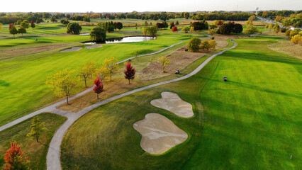 Aerial View of Scenic Golf Course in Autumn. Aerial view of a green golf course with sand bunkers, pathways, trees in fall colors, and a pond in the distance.
