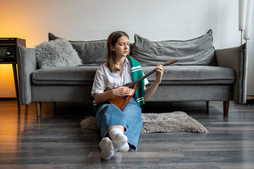 Relaxed woman musician strum balalaika strings, practice melodies, seated on living room floor beside sofa. Informal music session, song hobby with traditional stringed instrument, rhythm exploration