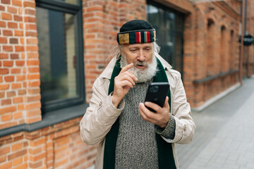 Portrait of homeless elderly man having smartphone conversation and raising index finger while...