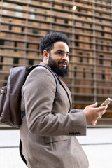 A fashionable African American businessman confidently strides through a vibrant modern urban landscape with a stylish backpack slung over one shoulder while engaging with his smartphone