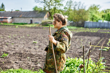Person in camouflage jacket holding wooden stick standing in field with green plants and trees in background rural setting farming gardening outdoor activity