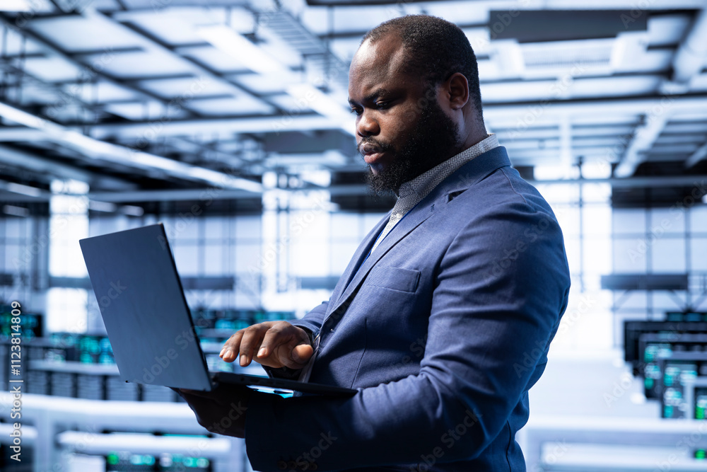 Poster engineer in server room establishment installing newly ordered storage units using laptop. african a