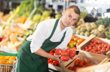 Friendly male seller in green apron enthusiastically offering fresh ripe tomatoes in organic grocery store filled with vibrant vegetables