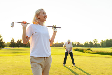 portrait of old woman golfer with club on golf course at sunset, elderly senior couple playing golf...
