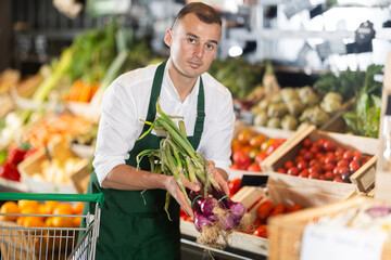Sales man works in vegetable store bio supermarket and goes through goods. He holds onion in hands, examines it, checks quality