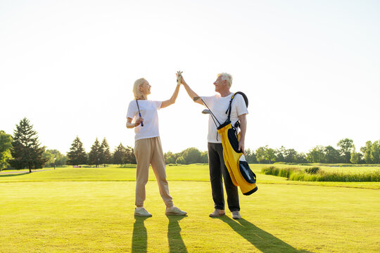 elderly senior couple in uniform celebrating victory and success in golf game and giving high five, old man and woman playing golf on golf course at sunset and doing outdoor sports
