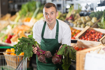 Smiling young adult vendor in green apron sorting fresh beets on display in farm market, surrounded by variety of colorful organic vegetables..