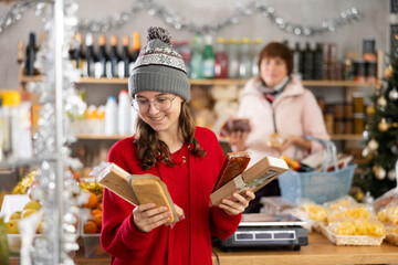 European girl purchaser choosing turrons in small grocery store decorated for New Year holiday with mother choosing food on shelves in background