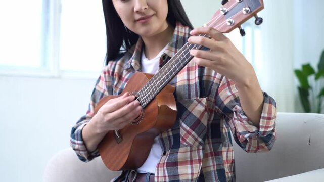 young woman playing ukulele at home and changing chords on strings close-up, female musician playing small acoustic guitar