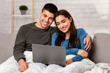 Fototapeta premium Stay At Home Concept. Self isolated spouses hugging and watching movie on laptop sitting on sofa, copy space