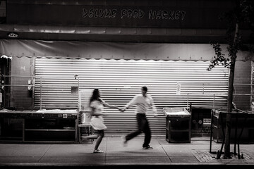 In a lively nighttime setting, a couple joyfully races past a closed food market, full of...