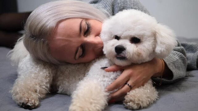 Blonde woman lies on grey blanket, gently hugging bichon frise dog with warmth and affection scene highlights their strong companionship, showing love and connection with her bichon frise dog. 
