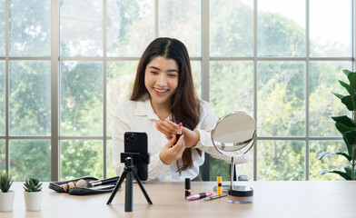 A woman filming her makeup tutorial in a bright room with large window, using a smartphone mounted...