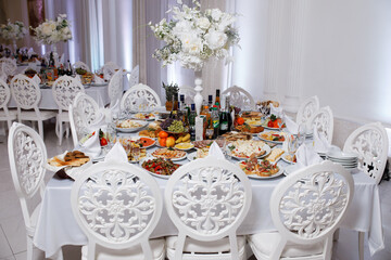 festively set table in a bright room. The table is round, covered with a white tablecloth and surrounded by white chairs with decorative carvings on the backs.