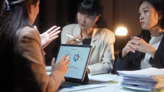 Three women are sitting around a table with a laptop open in front of them. They are discussing something on the screen, possibly related to business or work. The atmosphere seems to be serious
