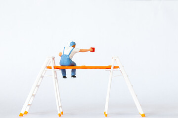 Miniature figure of a painter sitting on ladders holding a red paint can on white background