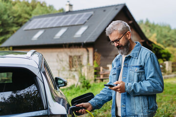 Man charging electric car in front of his house, plugging the charger into the charging port. House with solar panel system on roof behind him.