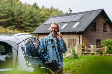Man charging electric car in front of his house while making phone call. House with solar panel...