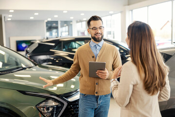 Smiling car seller with tablet explaining car performances to a customer at car showroom.