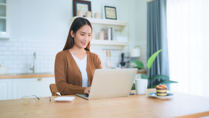 Beautiful young Asian woman working with laptop computer in home kitchen. Work at home