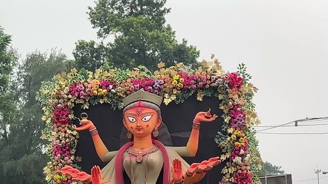Maa Durga idol on a small truck, with a black background, flower decorations, trees, greenery, and blue sky.