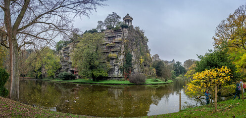 Naklejka premium Paris, France - 11 11 2024: Park Buttes Chaumont. View the central part of the park with footbridge, belvedere island, Temple of the Sibyl, the lake, remarkable trees and vegetation with fall colors.