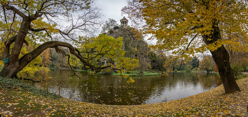 Paris, France - 11 16 2024: Park Buttes Chaumont. View the central part of the park with footbridge, belvedere island, Temple of the Sibyl, the lake, remarkable trees and vegetation with fall colors.