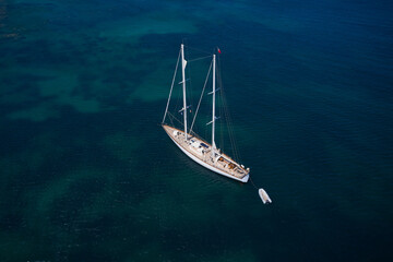 Large sailing yacht without sails anchored on clear blue water, aerial view. Sailing yacht with a dinghy visible on blue water, aerial view.