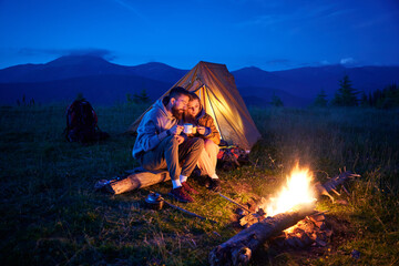 Couple hikers enjoys cozy moment by campfire, sipping hot drinks near glowing tent. Twilight sky and mountain backdrop create serene and romantic atmosphere in picturesque wilderness setting.