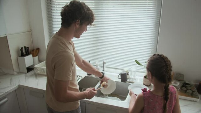 High angle view of cheerful Caucasian father giving daughter plates to wipe while teaching her doing housework at kitchen