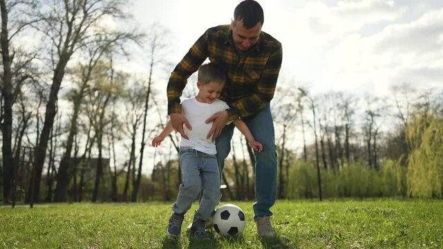 Father teaches son to play soccer. Happy family plays with ball outdoors. Father son bond builds confidence joy. Happy son father play ball together. Family enjoys playing ball outside on sunny day.