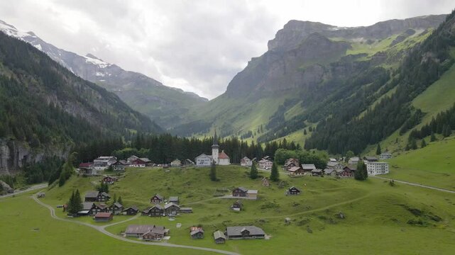 A serene village nestled in the Urnerboden valley, surrounded by lush green meadows, dense forests, and towering alpine mountains under a cloudy sky. Wide shot