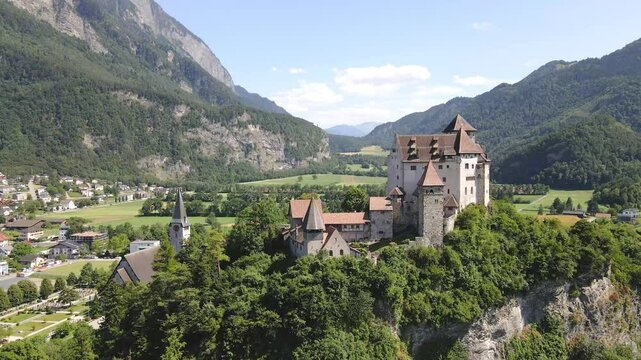 Aerial view of Burg Gutenburg in Balzers, Liechtenstein, surrounded by lush greenery and mountainous landscape under a clear blue sky, showcasing historical architecture and natural beauty. Wide shot.